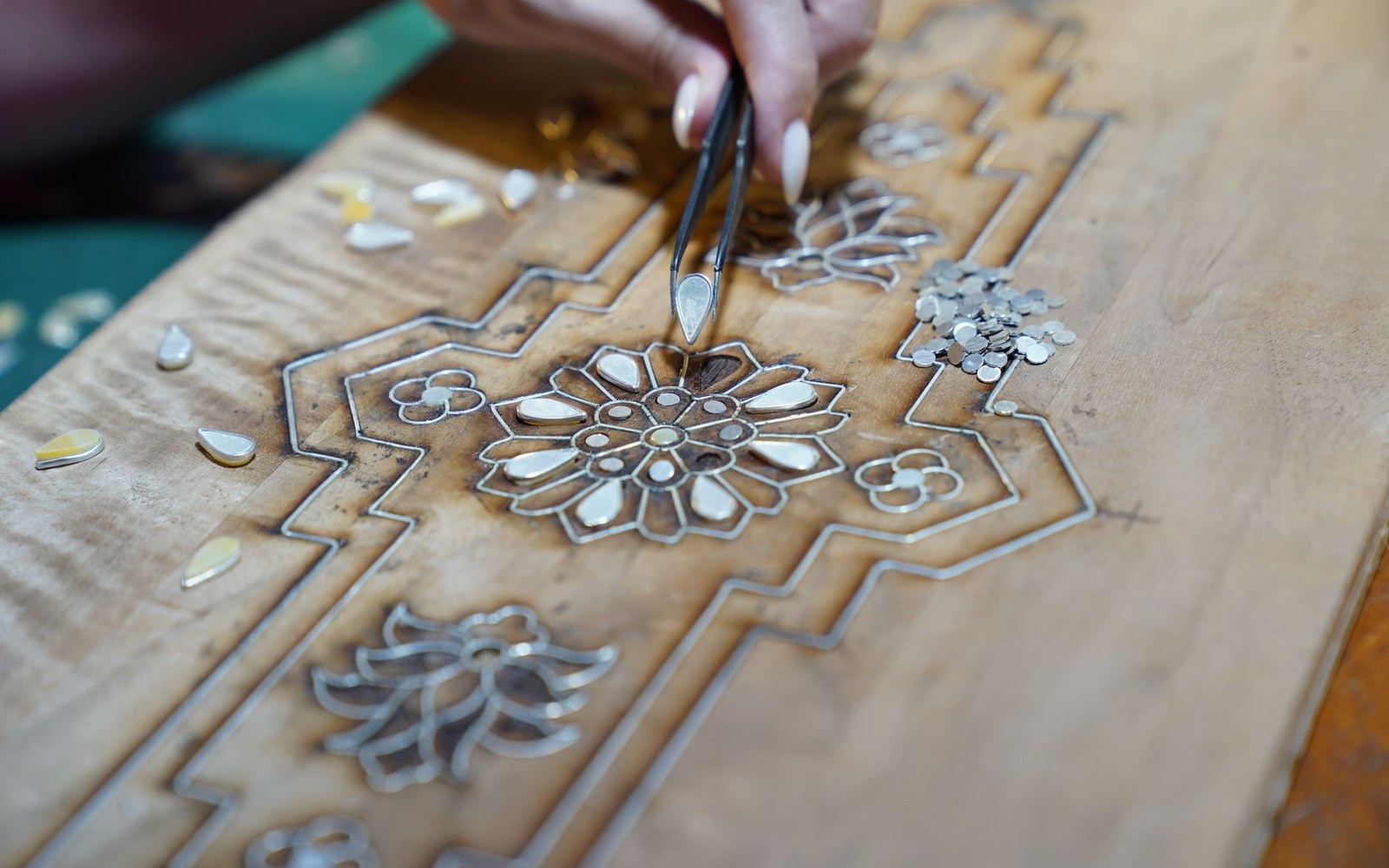 Craftsperson uses tweezers to place mother-of-pearl pieces into a carved wooden inlay pattern