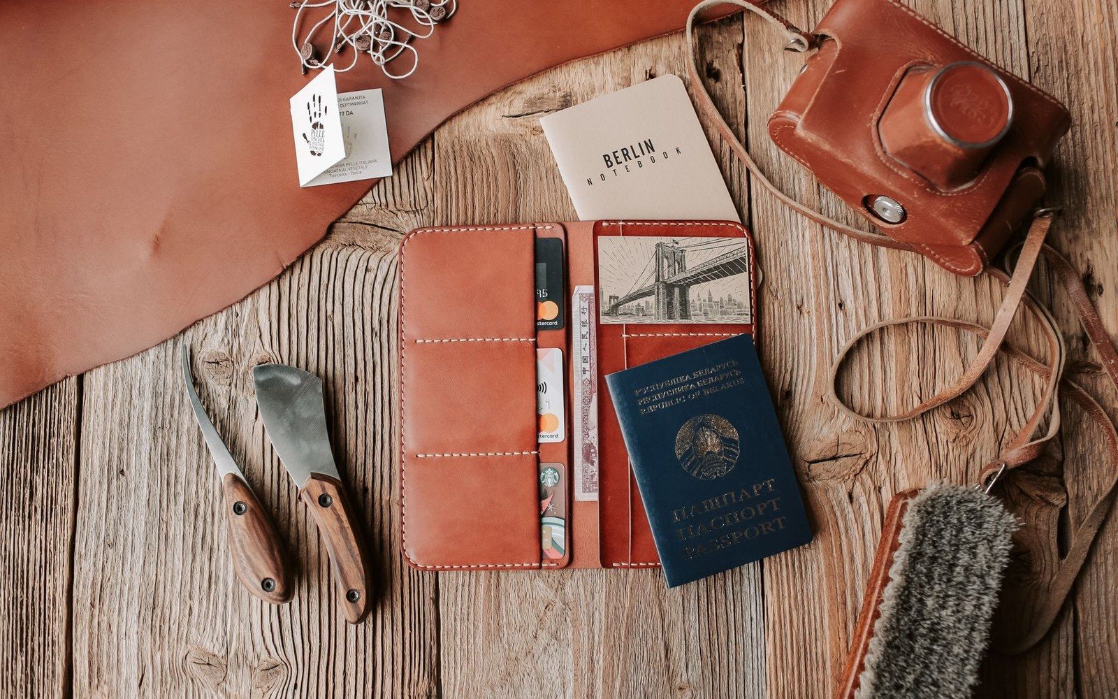 Leather travel wallet with passport and notebook beside a camera case and small knives on wood