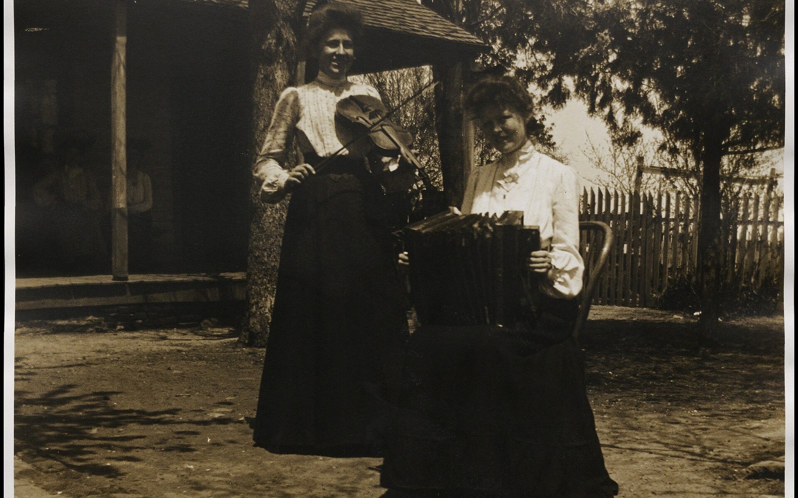 Two women in long skirts pose outdoors, one holding a violin, the other an accordion